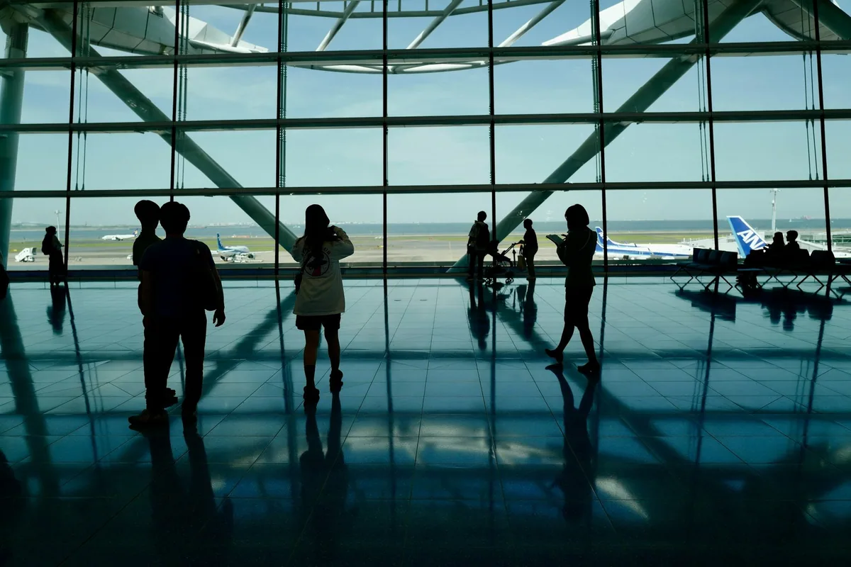 Families silhouetted against a tall terminal window, one adult carrying a small child, with aircraft on the apron beyond