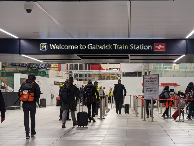 Passengers with wheeled suitcases walking in under the Welcome to Gatwick Train Station sign