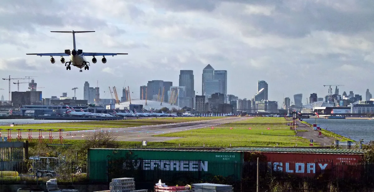 An airliner on final approach over the single runway at London City Airport, with the Canary Wharf towers on the skyline behind