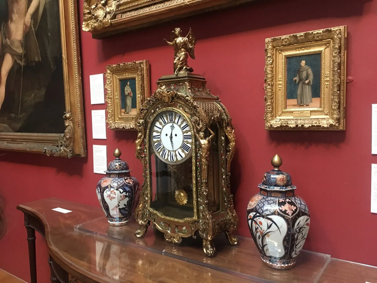 Gilt-framed old master paintings on a deep red wall above a gilded clock and two Imari jars