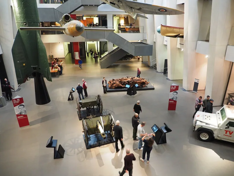 Visitors in the atrium of Imperial War Museum London among military vehicles, artillery and suspended aircraft