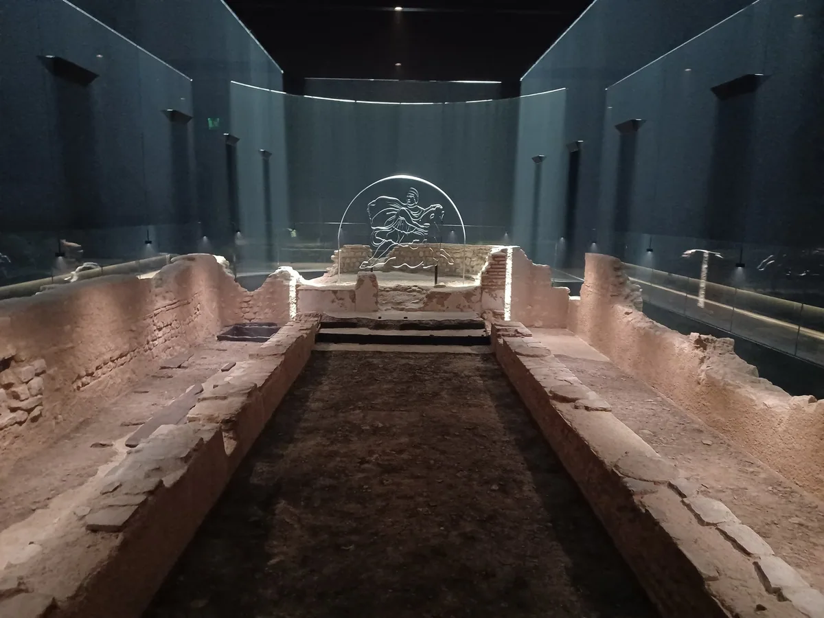 The reconstructed Roman temple of Mithras below street level, low stone walls lit in the dark with a Mithras outline at the far end