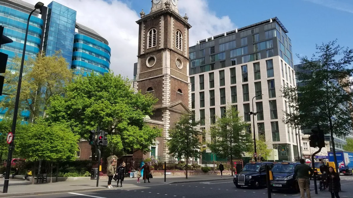 A brown-brick church tower rising between glass and stone office blocks in Aldgate, with street trees and traffic below