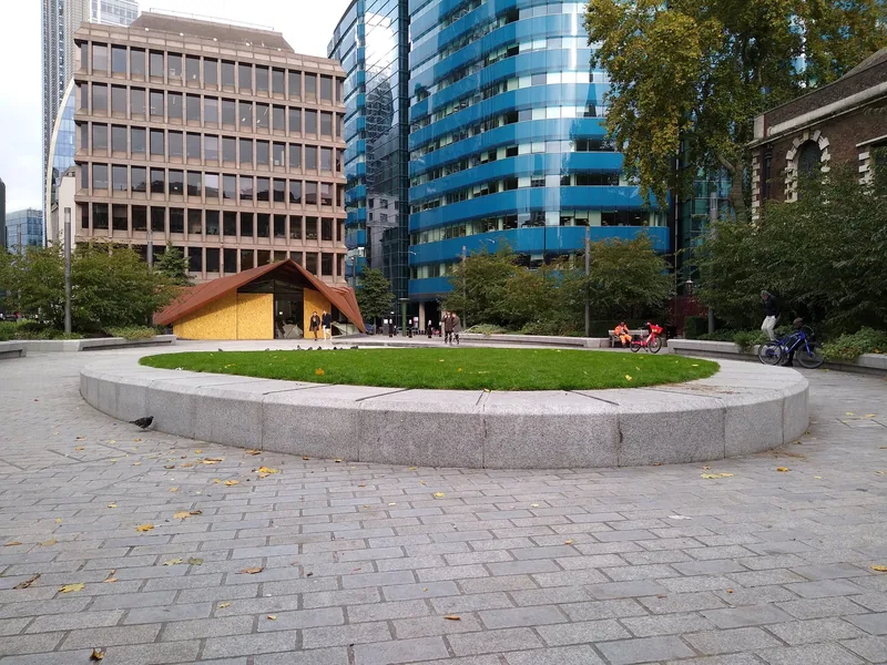 A raised circular lawn with a stone rim in Aldgate Square, a copper-roofed pavilion and City office blocks behind it