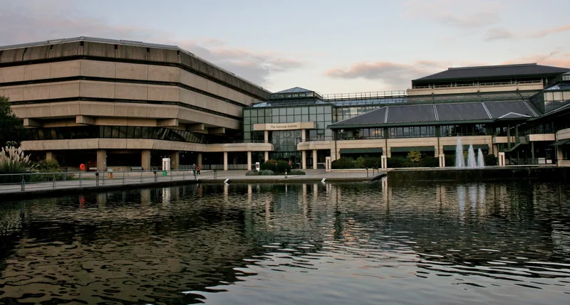 The stepped concrete 1970s wing of the National Archives at Kew beside the glass entrance building and the lake at dusk