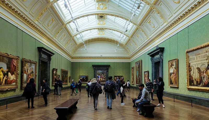 Visitors walking through a green-walled, skylit room of old master paintings inside the National Gallery