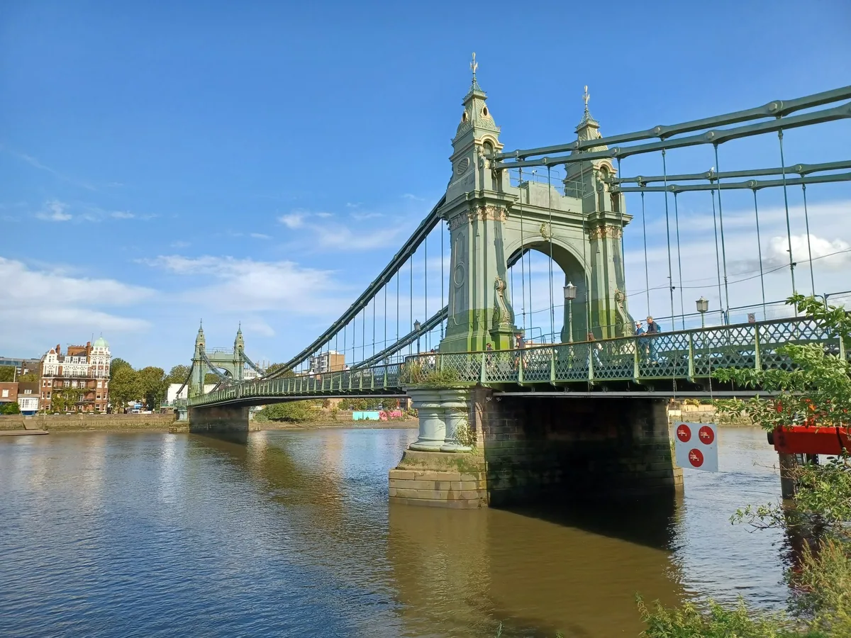 Hammersmith Bridge's green and gold suspension tower rising from its stone pier over the Thames, with a couple of people on the deck