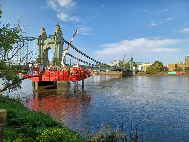 A red engineering platform with a crane standing in the Thames beside Hammersmith Bridge during repair work