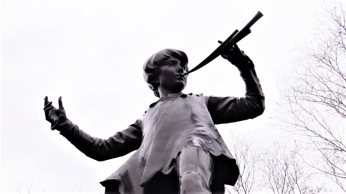 The bronze Peter Pan statue in Kensington Gardens seen from below, pipes raised to his lips and one hand outstretched against a pale sky