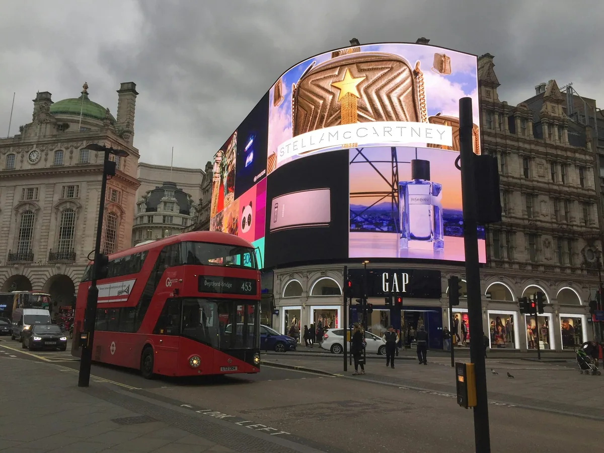 A red double-decker bus passing the curved illuminated screens of Piccadilly Circus at dusk