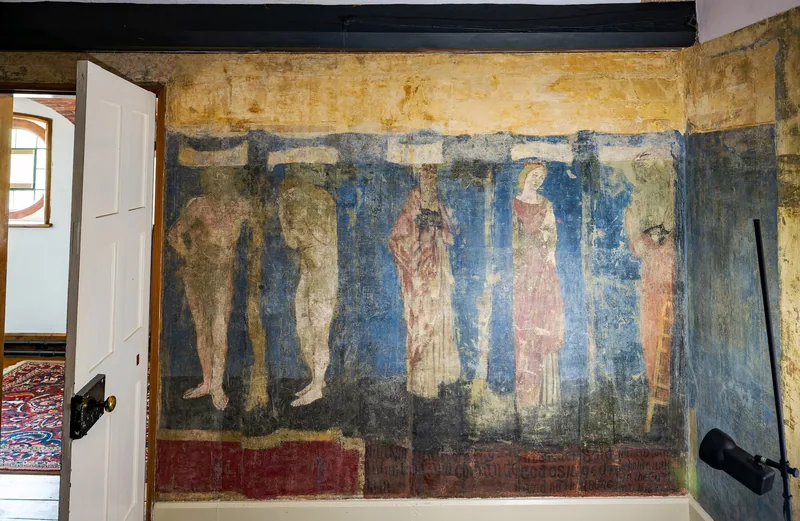 A faded painted wall of standing figures on a blue ground in a first-floor room at Red House, with an open panelled door beside it