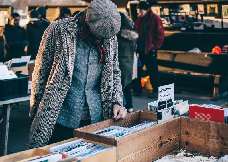A man in a tweed coat and flat cap leafing through a crate of second-hand books at the Southbank market stalls