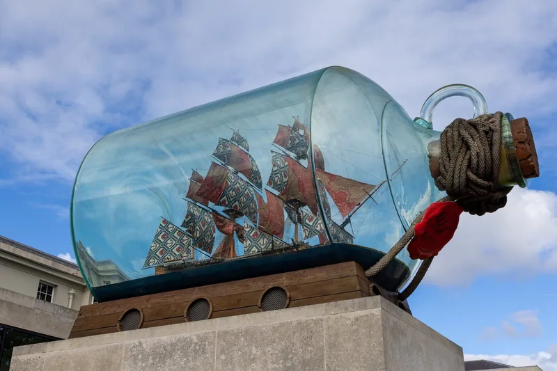 A giant model sailing ship with patterned fabric sails inside a glass bottle, mounted on a stone plinth at Greenwich