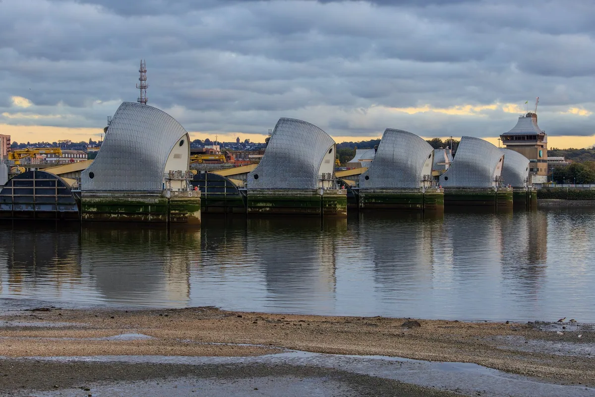 The steel piers of the Thames Barrier in a row across the river, reflected in calm water at low tide