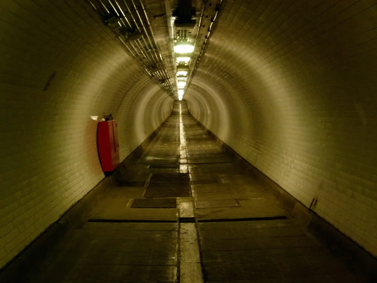 The white-tiled iron tube of the Woolwich foot tunnel, lit and empty, running away under the Thames