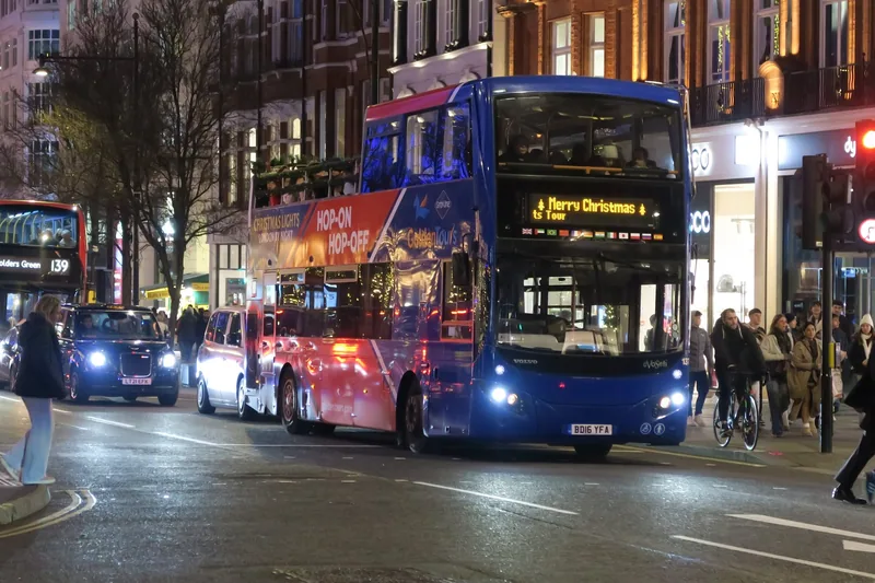 An open-top hop-on hop-off sightseeing bus in traffic on Oxford Street under the Christmas lights