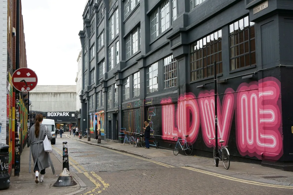 People walking along Ebor Street in Shoreditch past a large pink neon-style LOVE mural on a dark warehouse wall