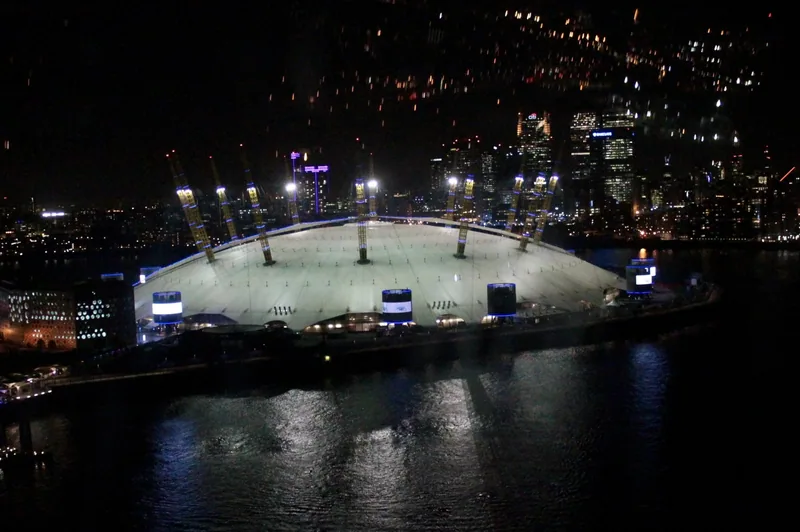 The lit O2 dome at night beside the Thames with Canary Wharf towers behind
