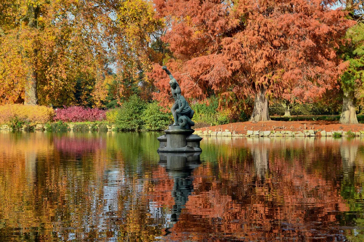 The Heracles and Achelous fountain standing in a Kew Gardens lake, ringed by orange and gold autumn trees mirrored in the water