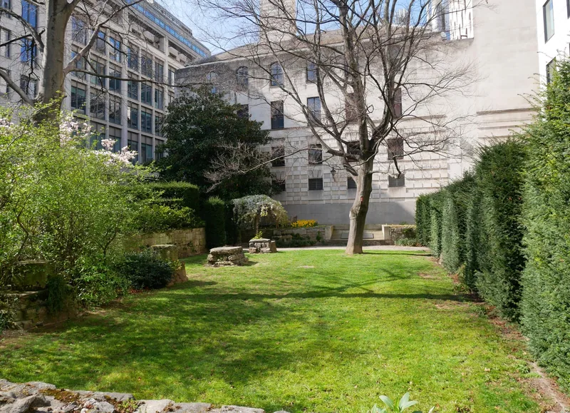 A small City lawn with the low stone footings of a vanished church set in the grass, hedged in by office buildings
