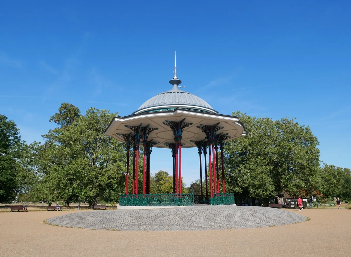 The ornate Victorian bandstand on Clapham Common, with its domed roof on slim painted columns, under a blue sky