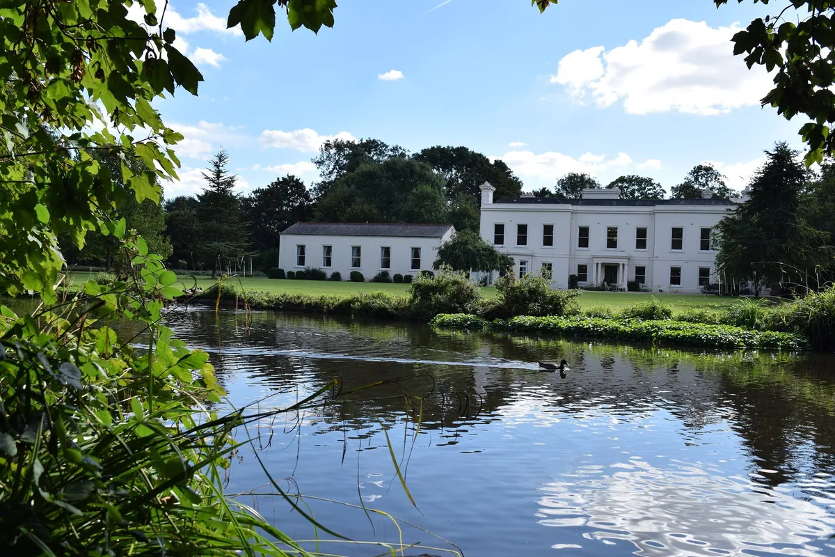 The white facade of Morden Hall seen across the River Wandle, with lawns and trees under a blue sky