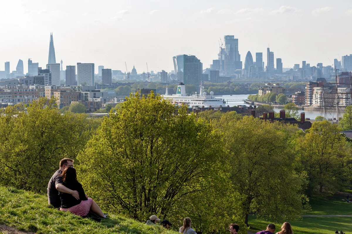 The City of London skyline seen over spring treetops from the hill in Greenwich Park, with a white ship moored on the Thames