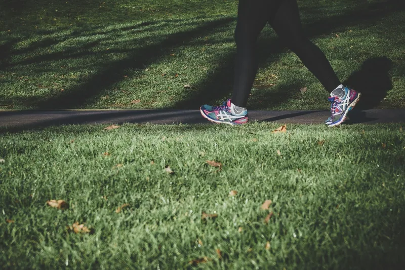 A runner in black leggings and bright trainers crossing a narrow tarmac path between areas of grass in a London park