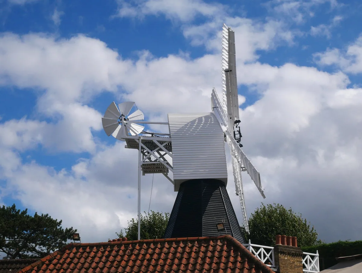 The white weatherboarded Wimbledon Windmill with its fantail and sails, standing above a tiled roof under broken cloud