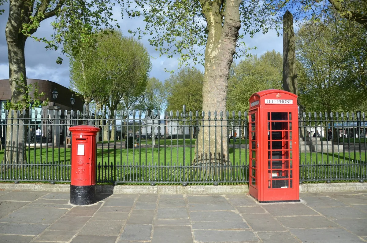 A red pillar post box and a red telephone box on a paved square beside park railings and trees