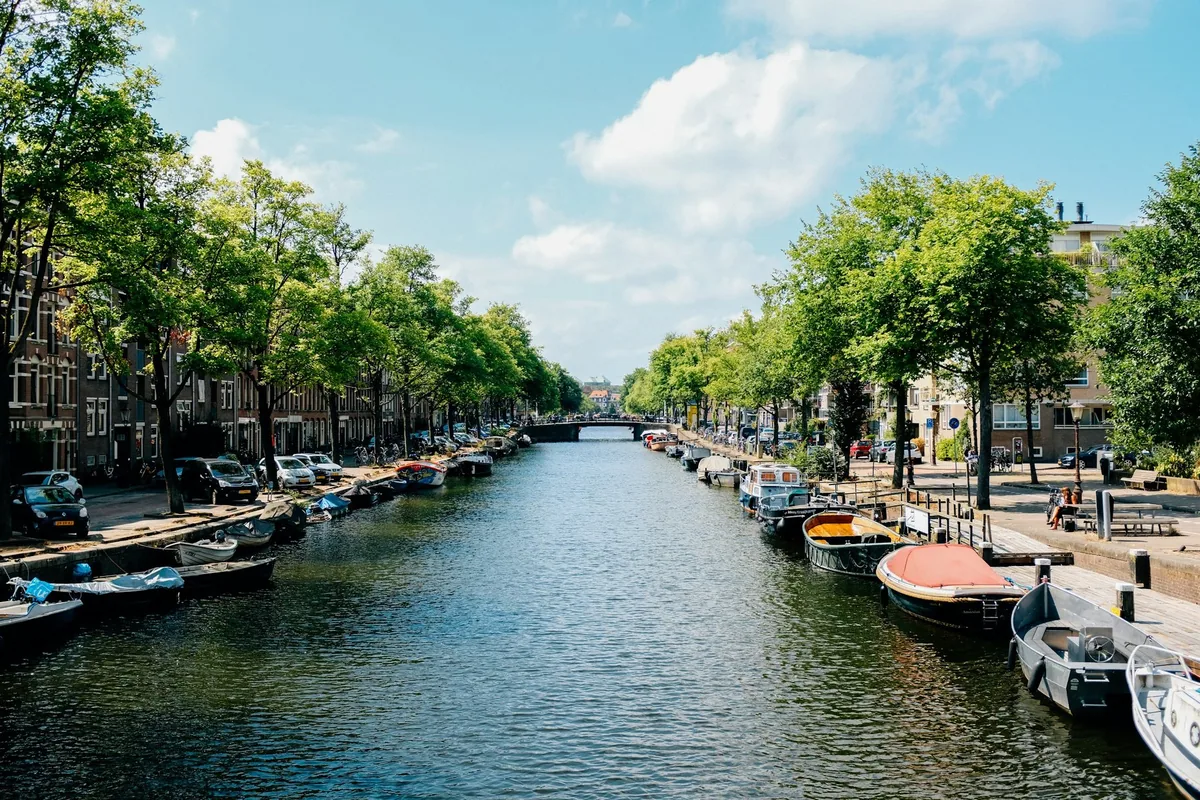 A tree-lined Amsterdam canal with small boats moored along both quays under a blue sky