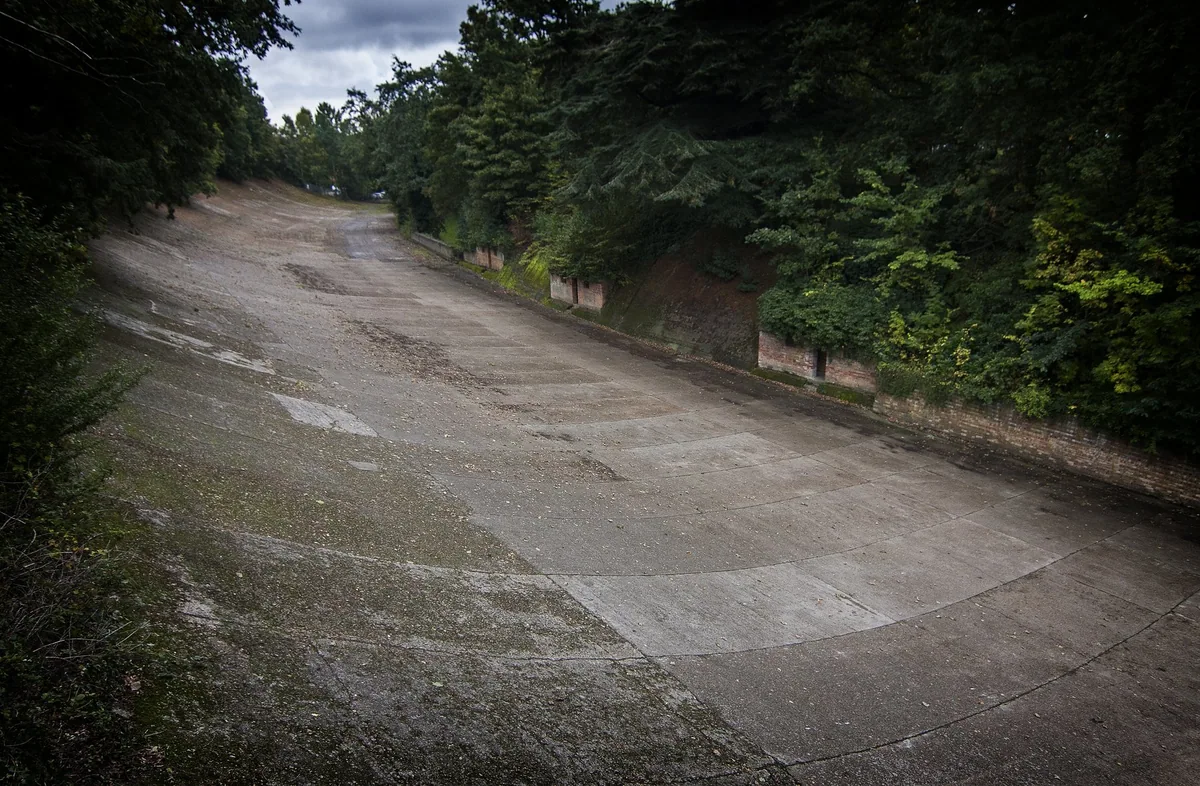 A surviving stretch of the steeply banked concrete racing track at Brooklands, cracked and empty, curving away between overgrown trees