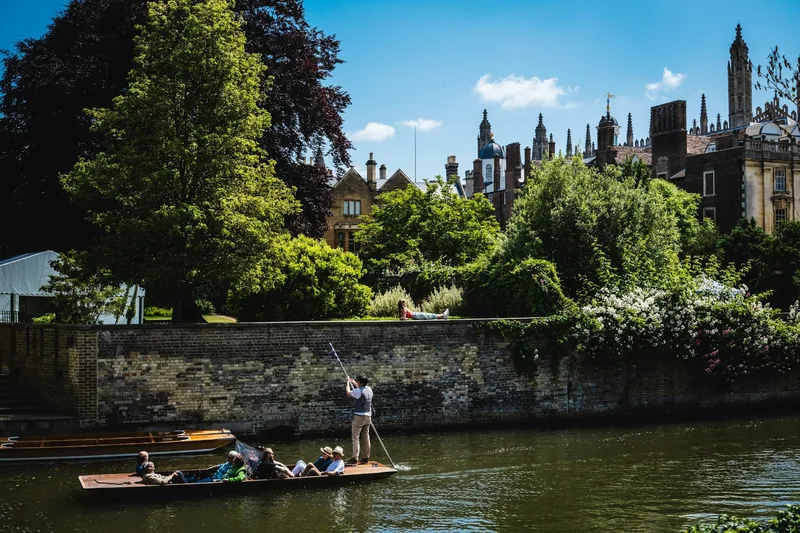 A punt being poled along the river Cam past college gardens and pinnacled roofs on a sunny day