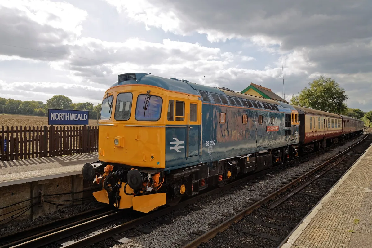 A blue and yellow Class 33 diesel with maroon and cream coaches at North Weald platform, the station nameboard on the left
