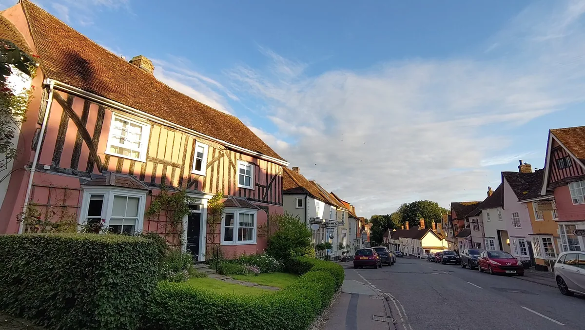 Pink-washed timber-framed houses leaning over Lavenham High Street in evening light, with colour-washed cottages down the street beyond