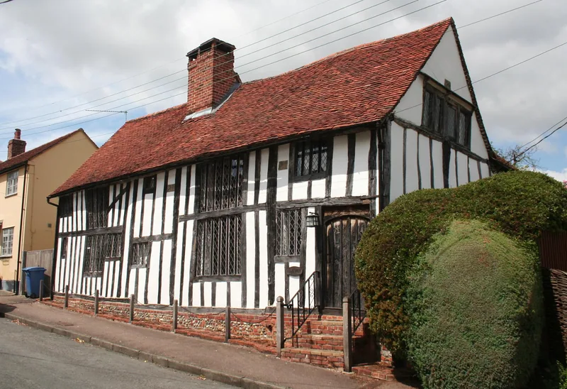 A black-and-white timber-framed hall house in Lavenham with a steep tiled roof, leaded windows and a clipped hedge beside the door
