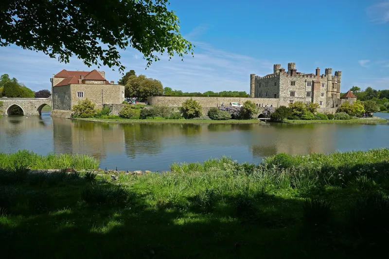 Leeds Castle and its stone bridge seen across the lake from the far bank on a sunny day