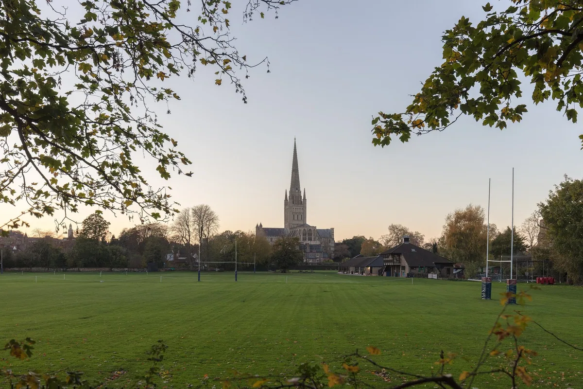 The spire of Norwich Cathedral rising above the rooftops and trees at dusk, seen across a wide green playing field