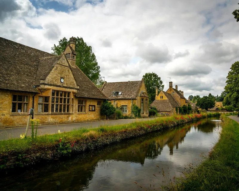 A Cotswolds village lane running beside a slow stream, with stone houses and mossy roofs