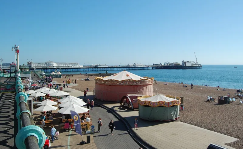Brighton seafront promenade with cafe parasols and a carousel tent, the shingle beach and pier beyond