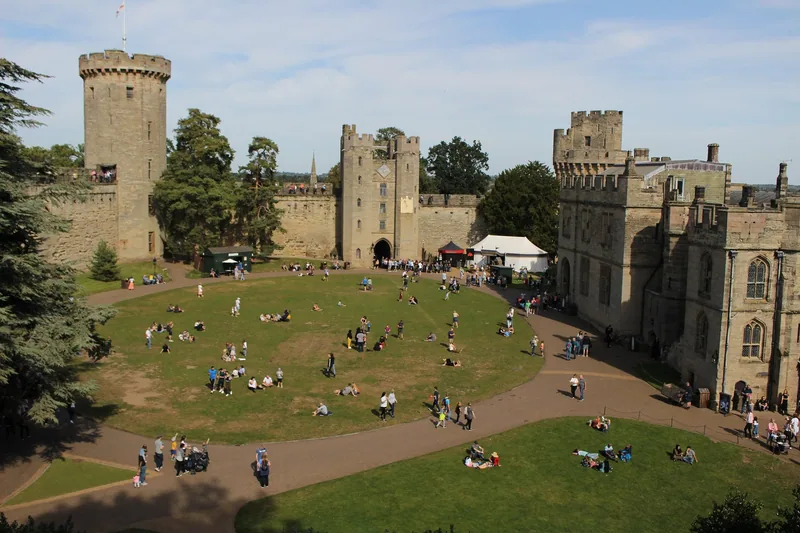Looking down on the grassy courtyard of Warwick Castle, with visitors sitting on the lawn below the towers