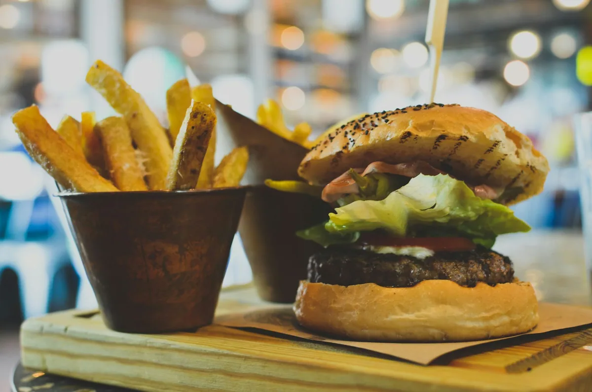 A burger with lettuce, tomato and a sesame bun on a wooden board beside a metal cup of thick-cut chips