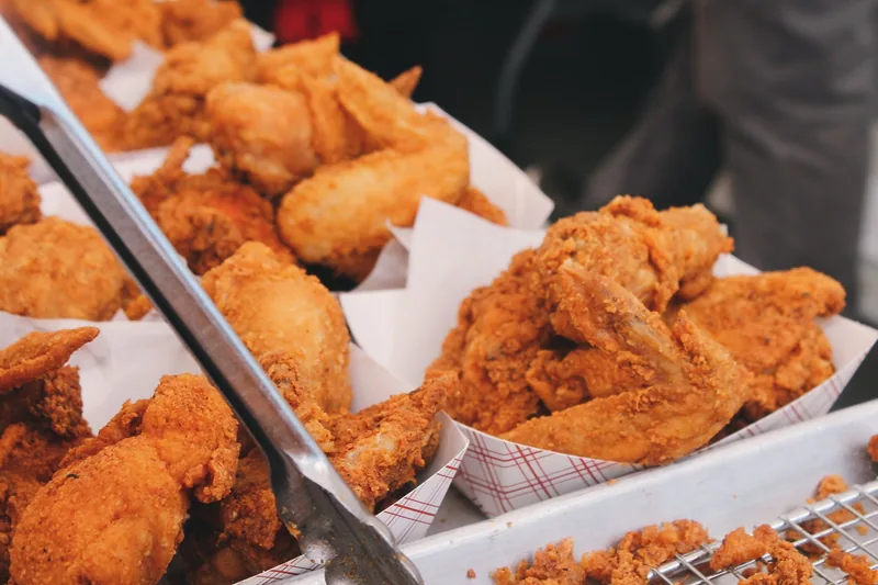 Crisp fried chicken wings and drumsticks piled in paper trays at a takeaway counter, with serving tongs