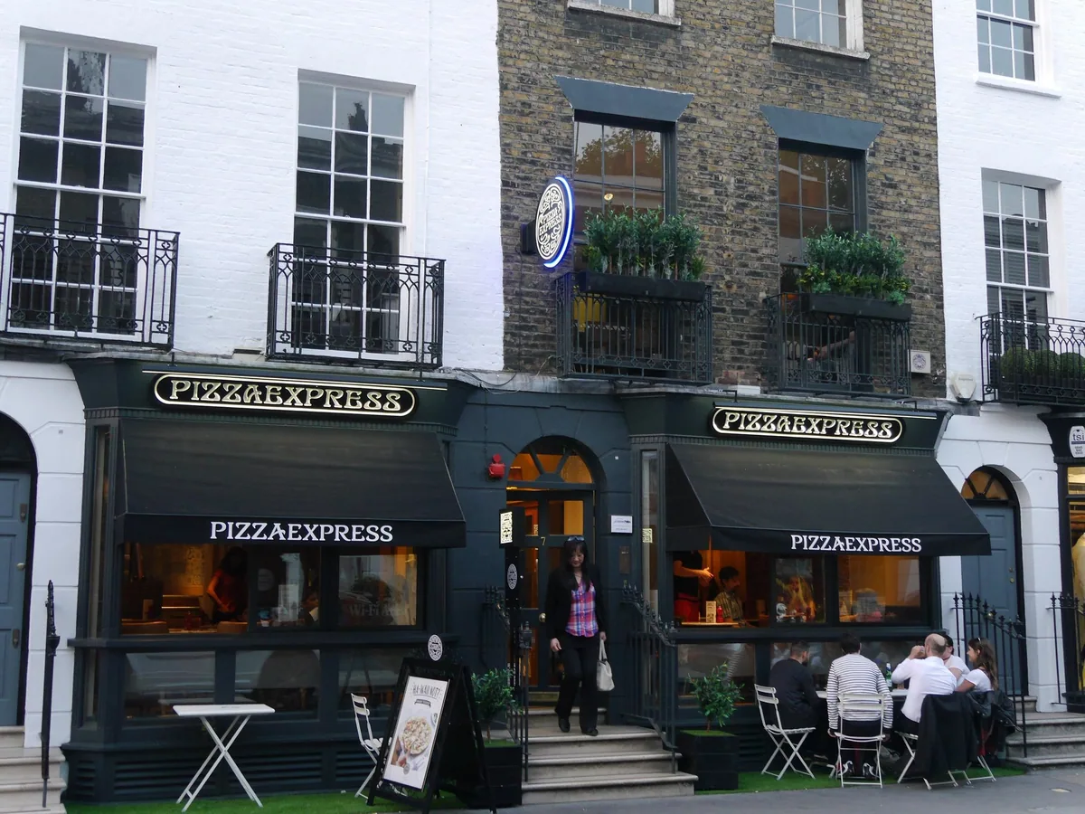 The dark green frontage of a Pizza Express branch on Beauchamp Place in London, with pavement tables