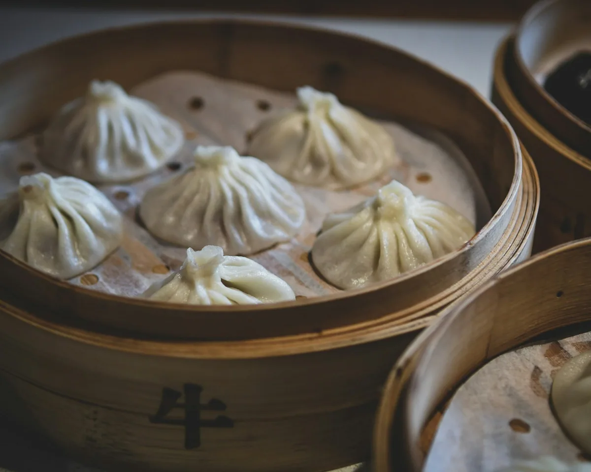 Hand-pleated soup dumplings resting in a round bamboo steamer, with more baskets stacked alongside
