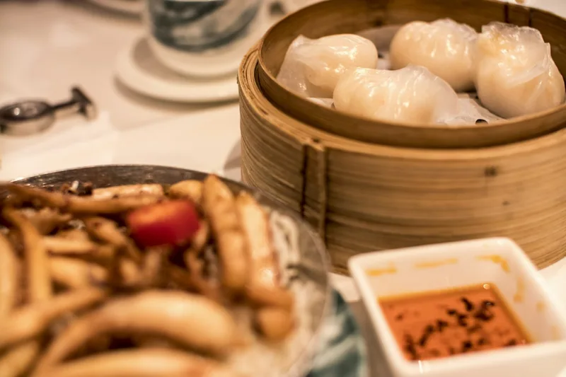 Translucent prawn dumplings in a bamboo steamer on a laid restaurant table, with a dish of chilli sauce