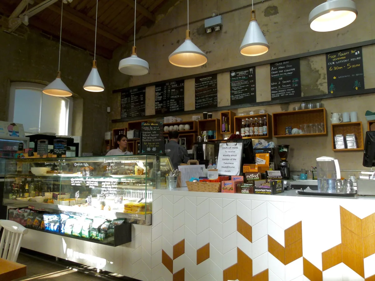 The order counter of a London cafe, with chalkboard menus above and a glass cabinet of cakes and sandwiches