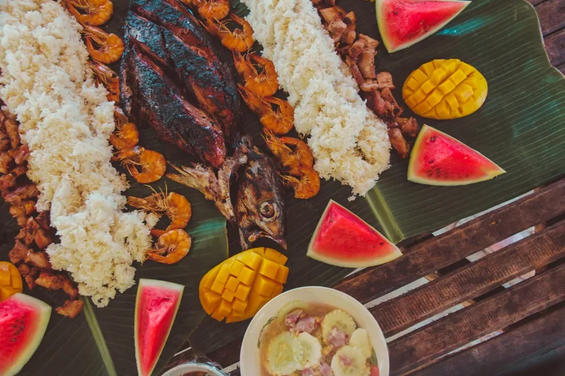 A shared feast laid out on banana leaves: grilled fish, prawns, rice, pork, mango and watermelon