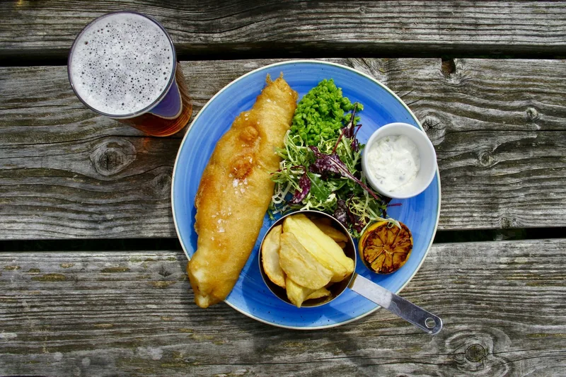 Battered fish with chips, salad and tartare sauce beside a pint of ale on a wooden pub table