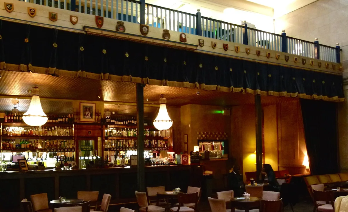 The galleried interior of a London pub, with a long bar, chandeliers and a balcony under a high ceiling